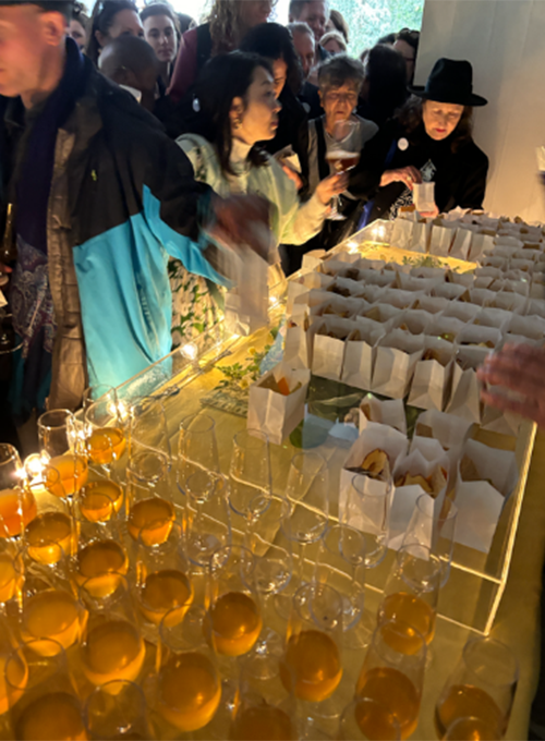 A large audience in front of a pile of glasses and paper bags lined up on a table