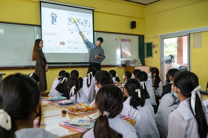 A teacher giving a lecture in a classroom while pointing at a screen with a projector on it, and students listening to the lecture