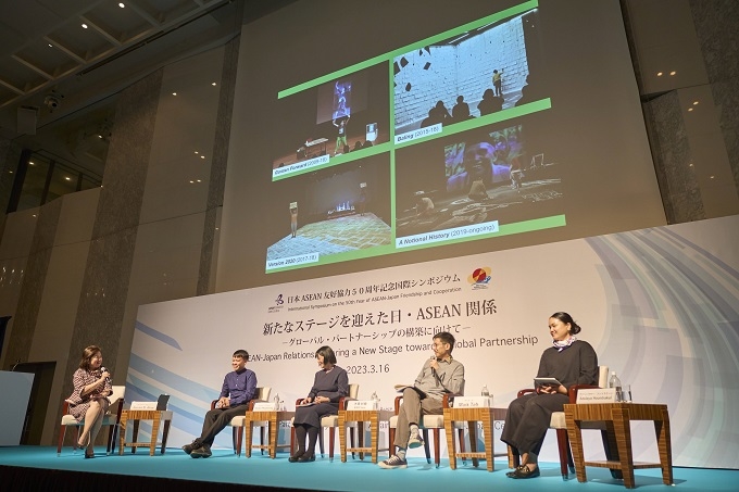 Four experts and the moderator were seated on chairs on a stage set up at the front of the large venue, holding a symposium. In the background was the society's signboard, and above it was a large screen projecting images.