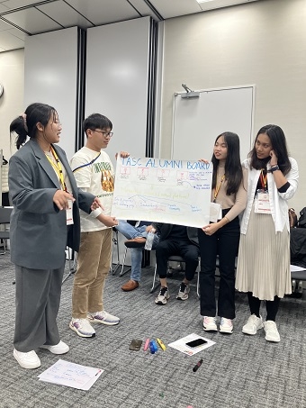 In one corner of the room, a man and a woman are holding a piece of paper with 'TASC ALUMNI BOARD' written on it, and two women are standing on either side of it, having a conversation.